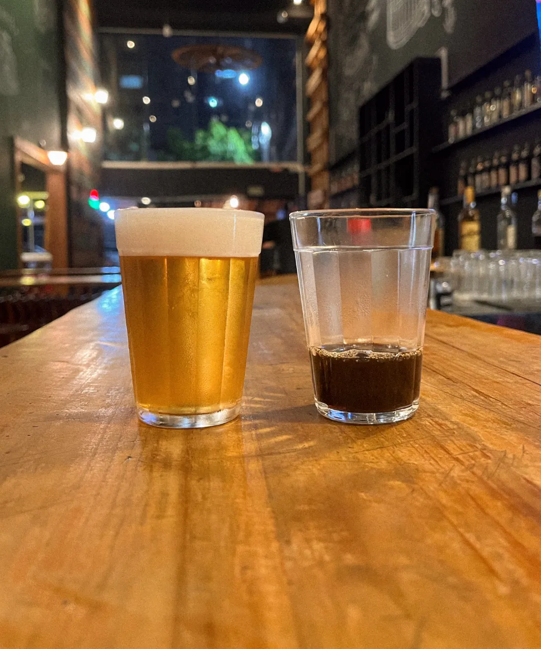Copo de chopp e copo americano com café sobre o balcão de madeira do Barile, com o salão ao fundo