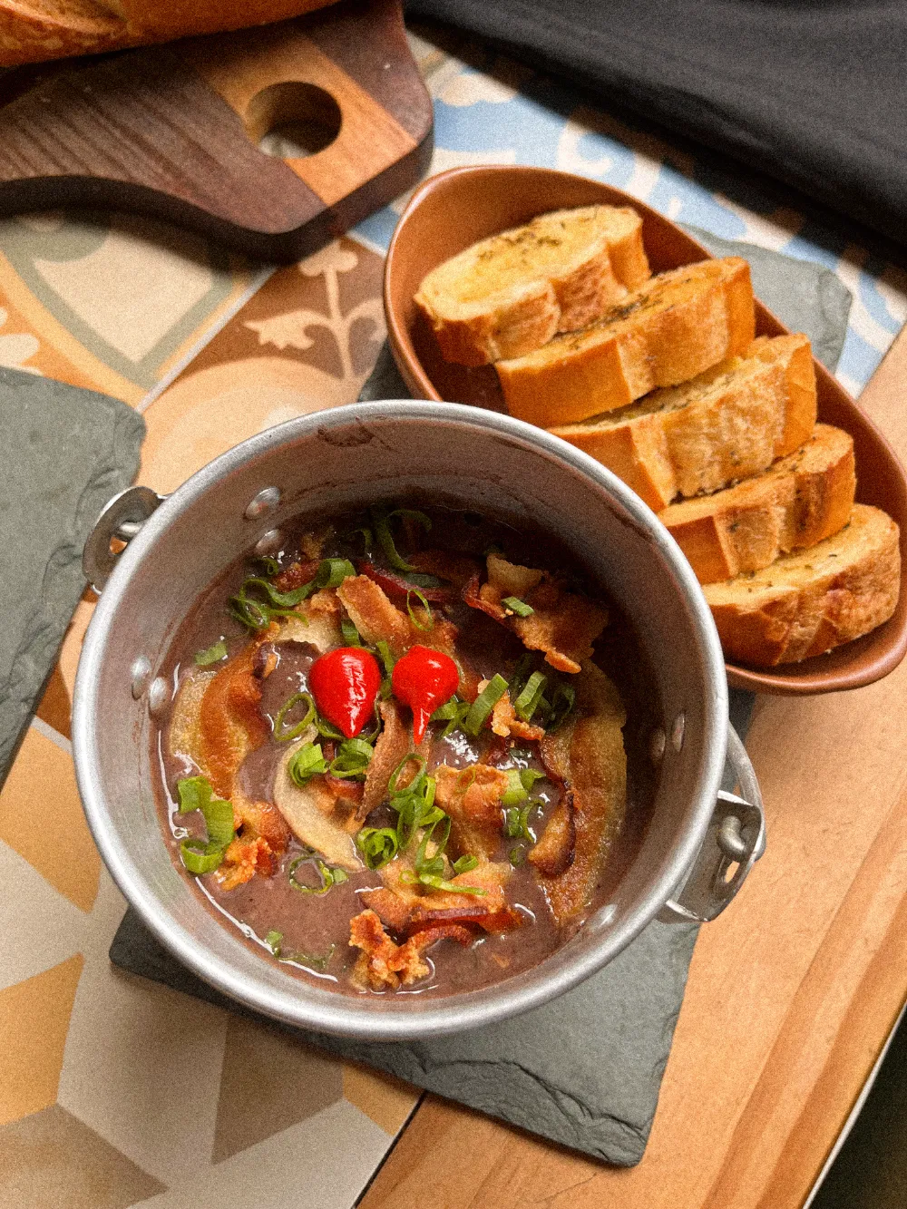 Caldo servido em panelinha de alumínio com bacon e pimenta, ao lado de torradas