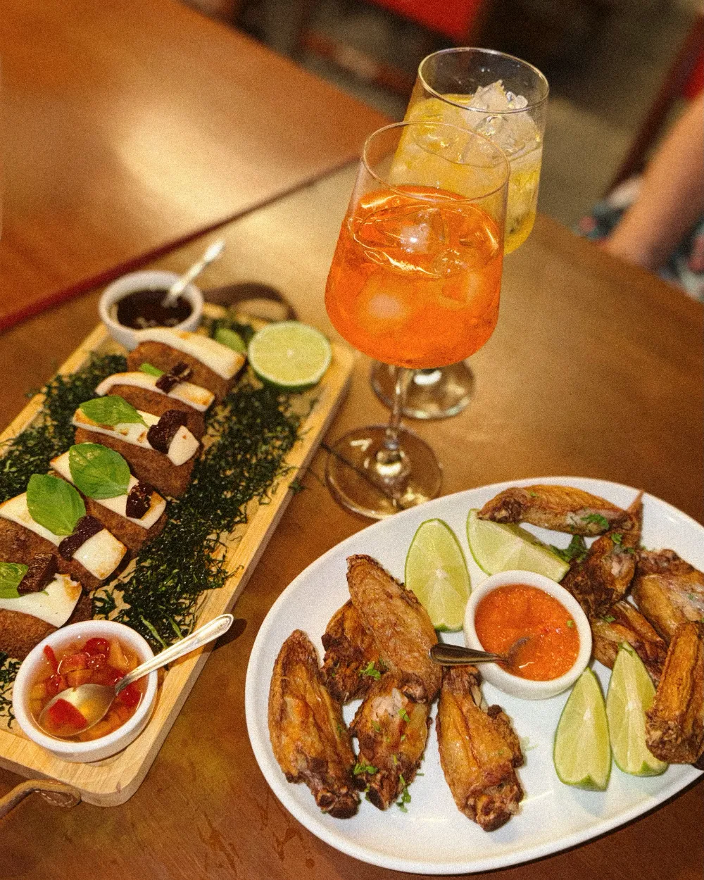 Tábua de carne com queijo, prato de asinhas de frango com molho e limão e dois drinks sobre a mesa de madeira